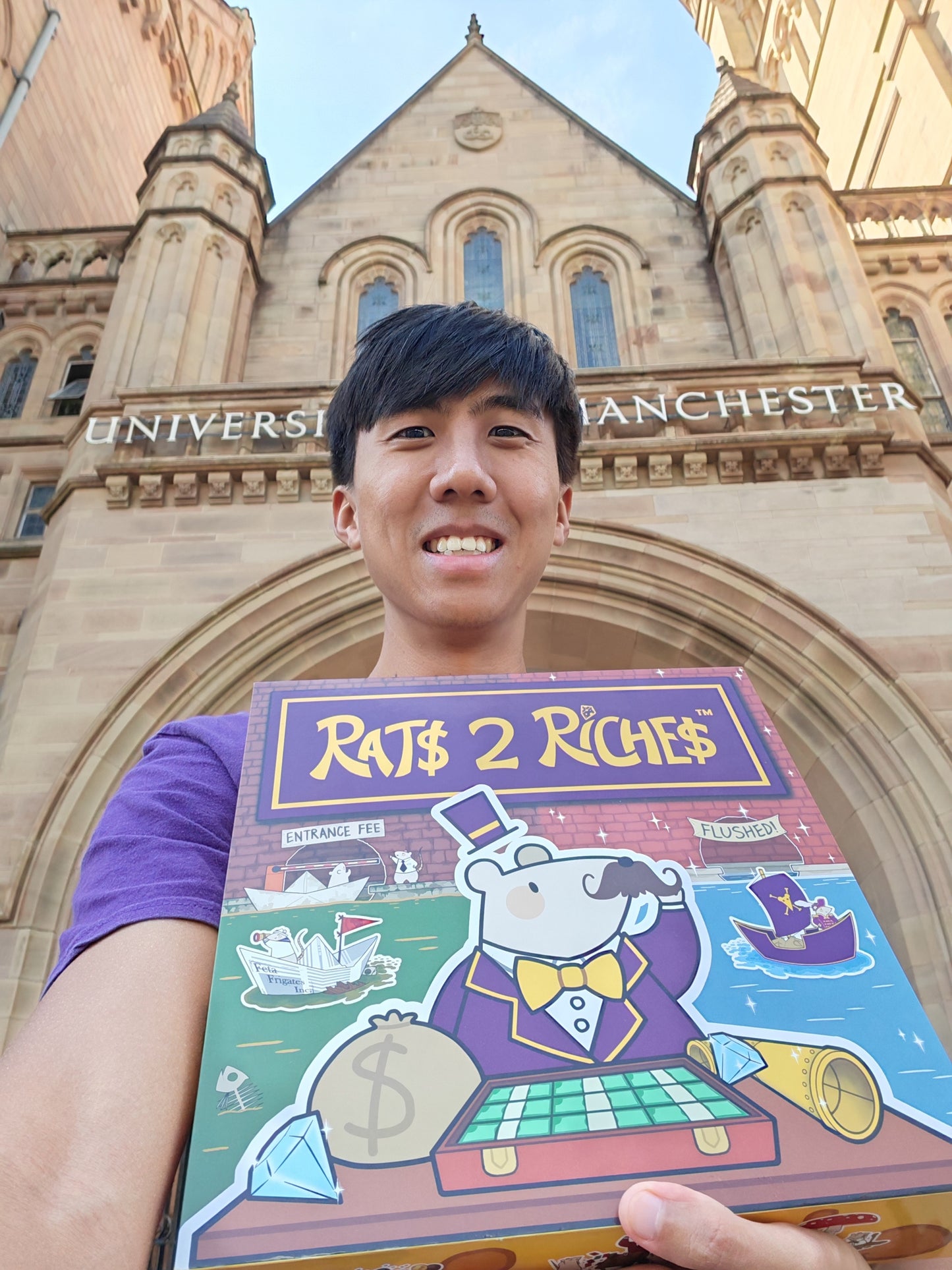 Person holding a 'Rats 2 Riches' board game box in front of a University of Manchester building.