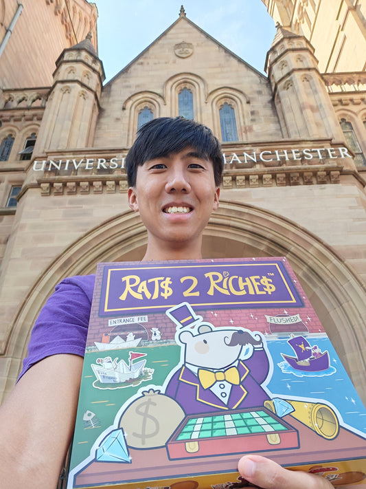 Person holding a 'Rats 2 Riches' board game box in front of a University of Manchester building.